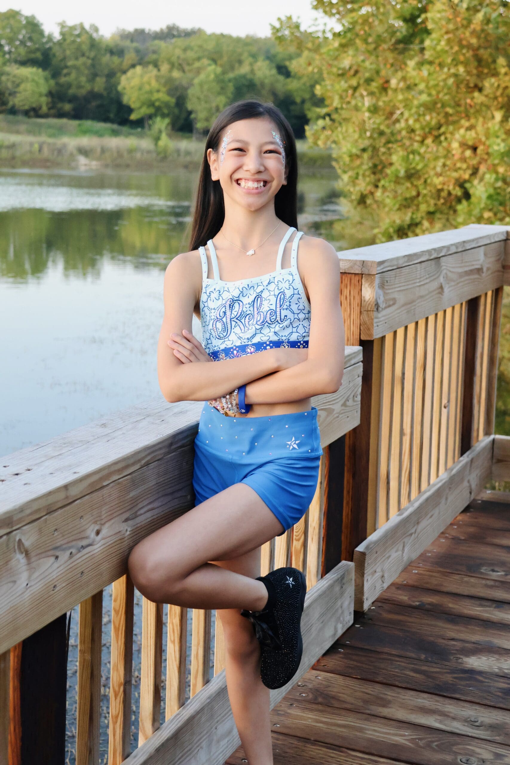 Roselyn leaning on a wooden railing by a lake, wearing a blue Rebel Athletics set, smiling with trees and water in the background.