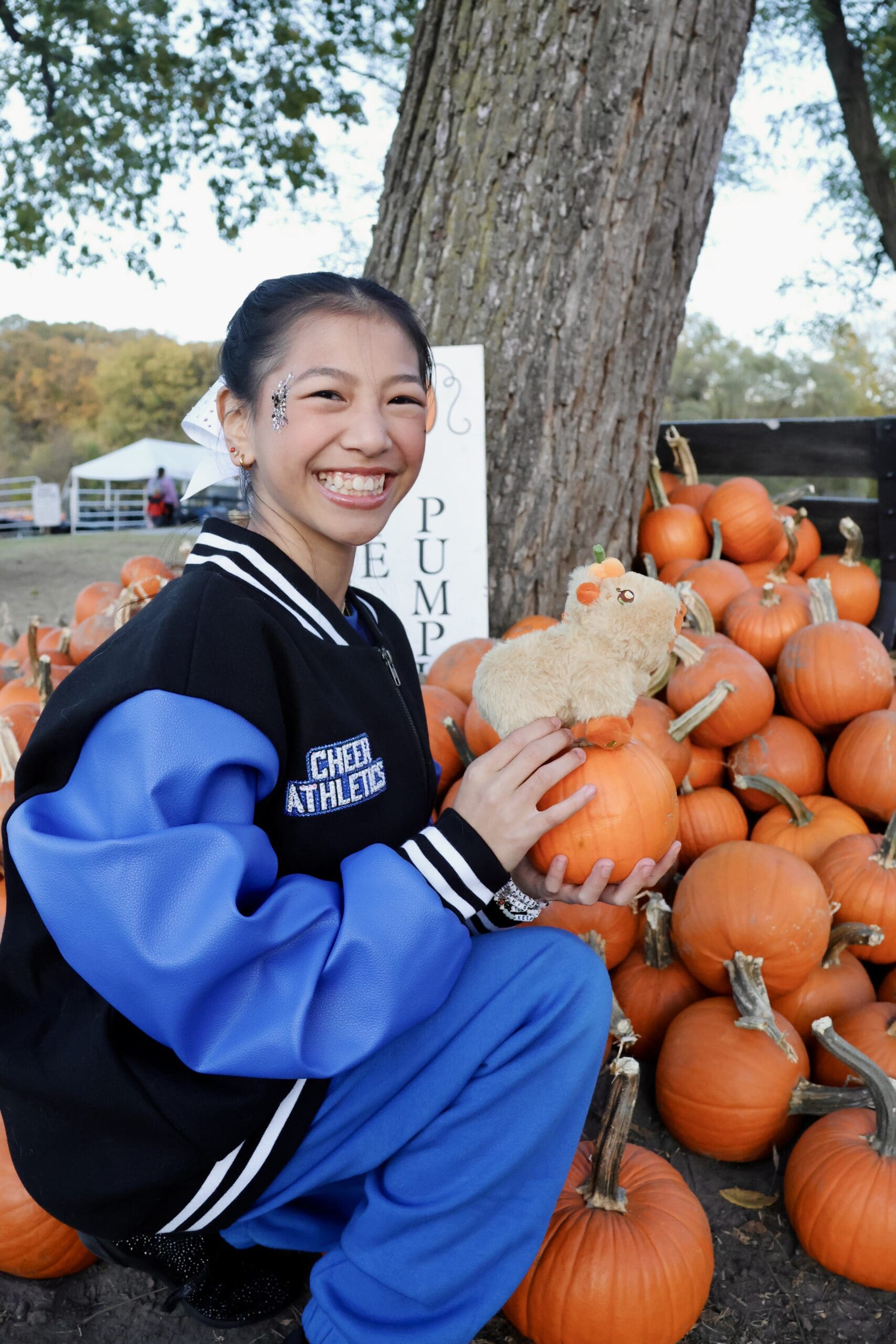 Roselyn sitting outdoors by a pile of bright orange pumpkins, wearing a Rebel Athletics letterman jacket and smiling during fall season.